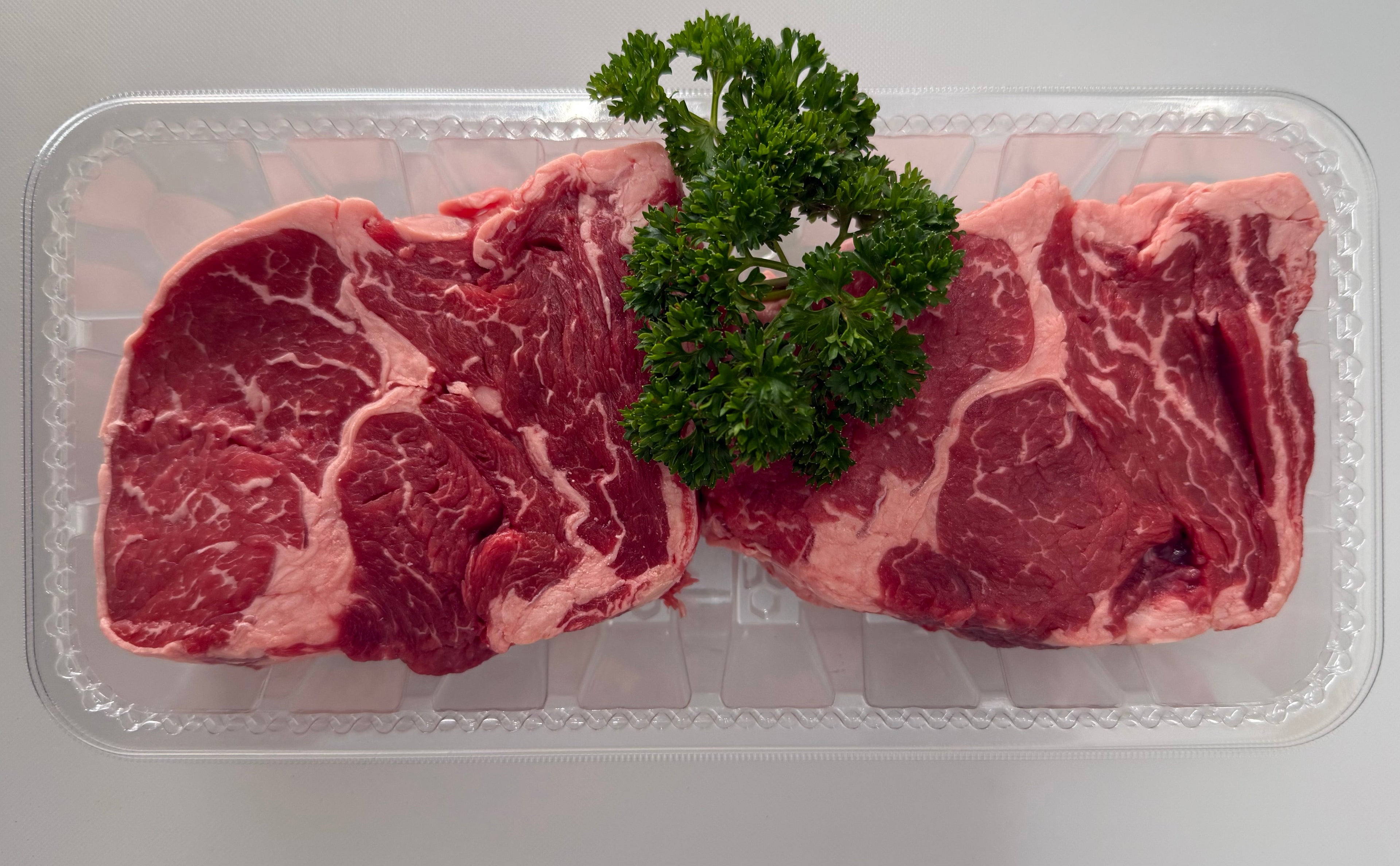 Two raw steaks on a clear plastic tray with fresh parsley on a white background