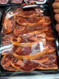 A black plastic tray filled with lamb chops, covered with clear plastic wrap, photographed in a grocery or butcher setting. 
