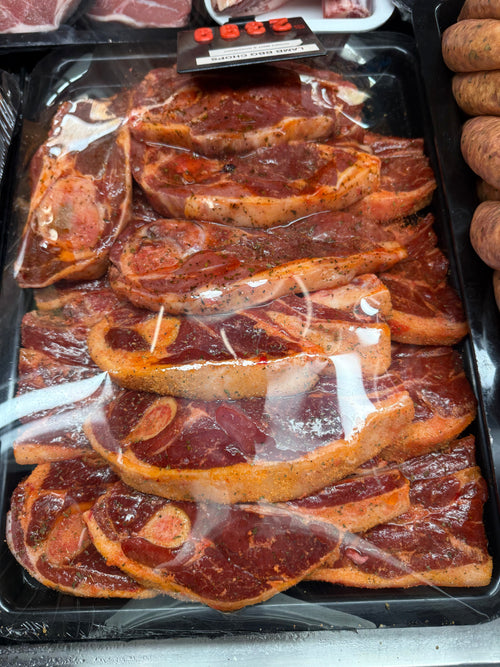 A black plastic tray filled with lamb chops, covered with clear plastic wrap, photographed in a grocery or butcher setting. 