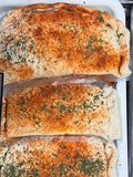 Three stuffed calzones topped with a golden-brown seasoning mix and chopped green herbs, arranged in a white baking tray next to a dark tray containing sliced red meat.