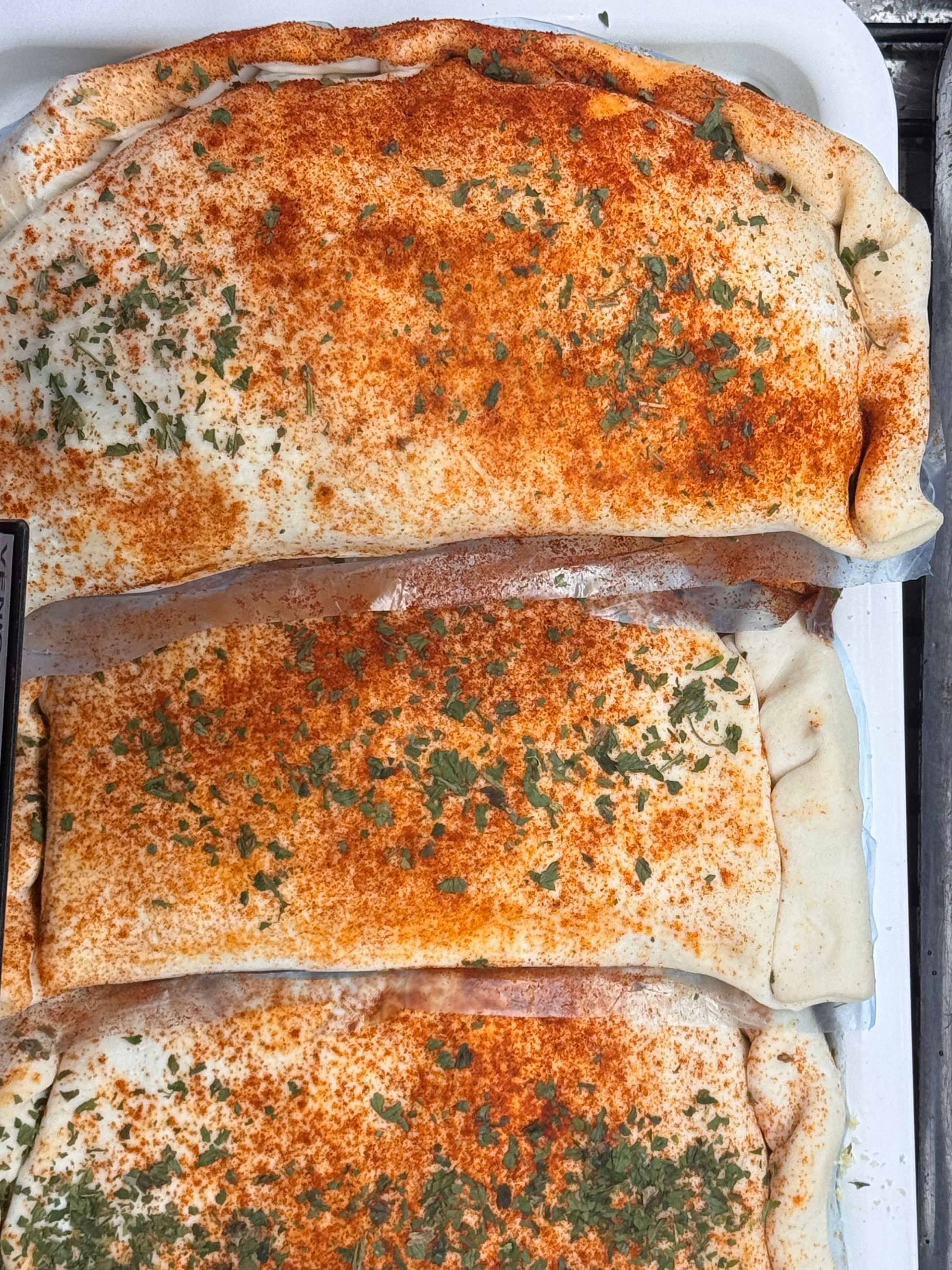 Three stuffed calzones topped with a golden-brown seasoning mix and chopped green herbs, arranged in a white baking tray next to a dark tray containing sliced red meat.